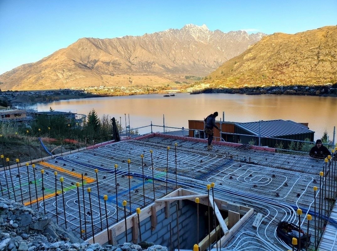 Hydronic underfloor heating loops laid over reinforcing mesh before a concrete slab pour, with a lake and mountains behind